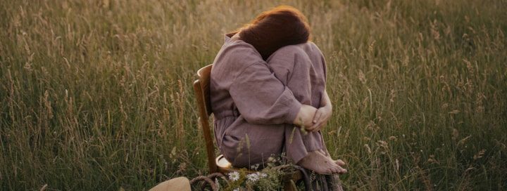 Eine Person sitzt alleinim hohen Gras, die Knie an den Körper gezogen. Das Bild vermittelt stille Verletzlichkeit und den Wunsch nach Schutz